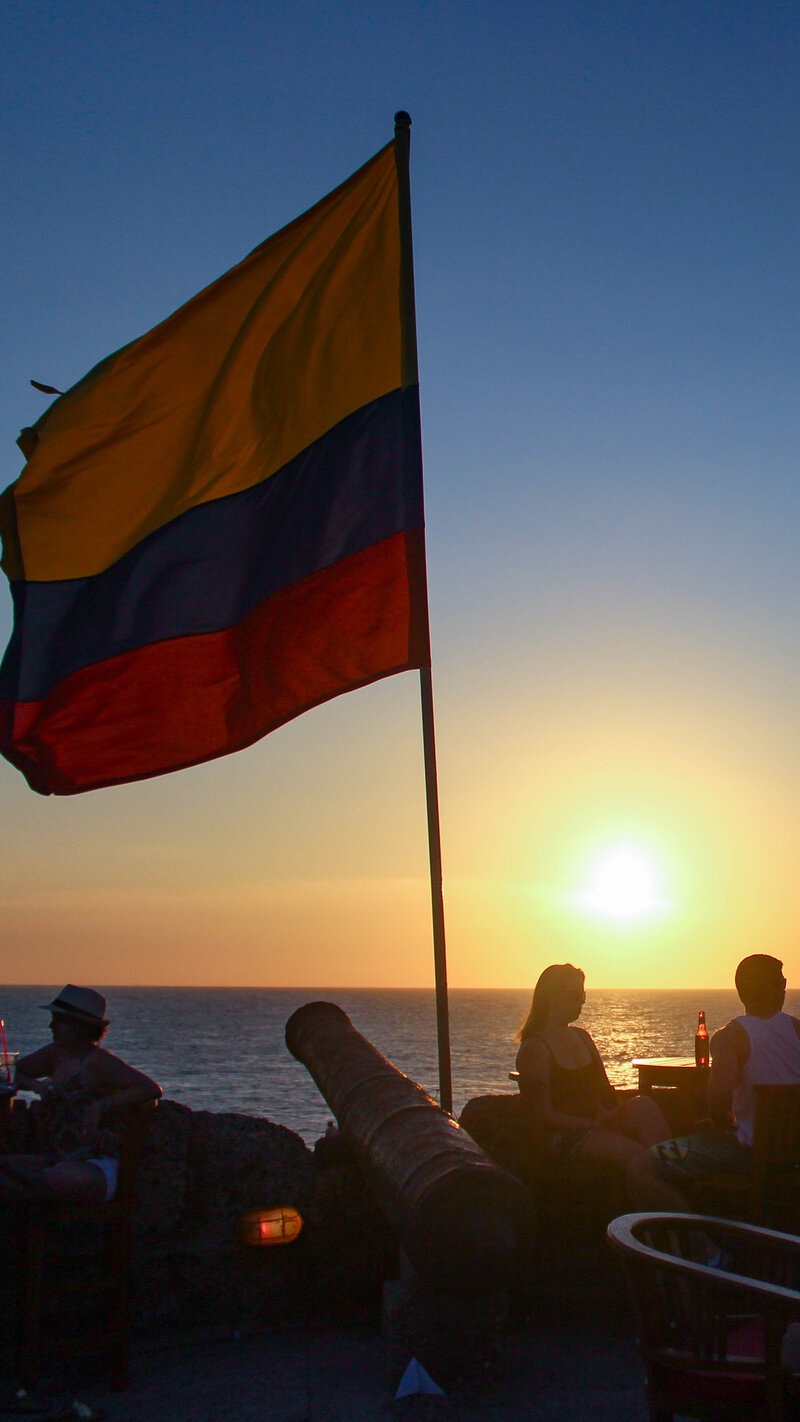 Silhouettes of people enjoying drinks at sunset on the walls of Cartagena, with a Colombian flag waving in the breeze and an old cannon pointing toward the Caribbean Sea ©Stephanie Dosch | theViatrix Curated Colombia Itinerary