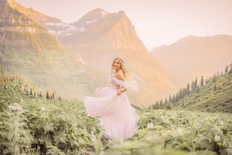 senior girl with large hair wearing a dress while standing in a field of wild flowers with mountains in the background