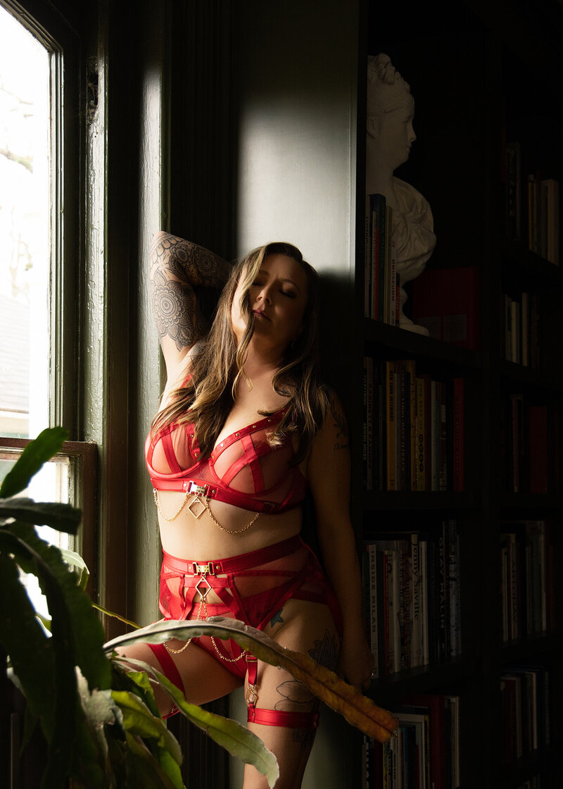 Woman in red lingerie standing confidently by a window and bookshelf, photographed during a boudoir session in an Atlanta luxury home by Christie Newell