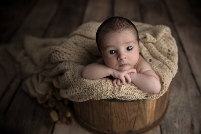 Newborn boy in beige wrap, alert and curious, posed in a rustic bucket for a timeless fine-art portrait.