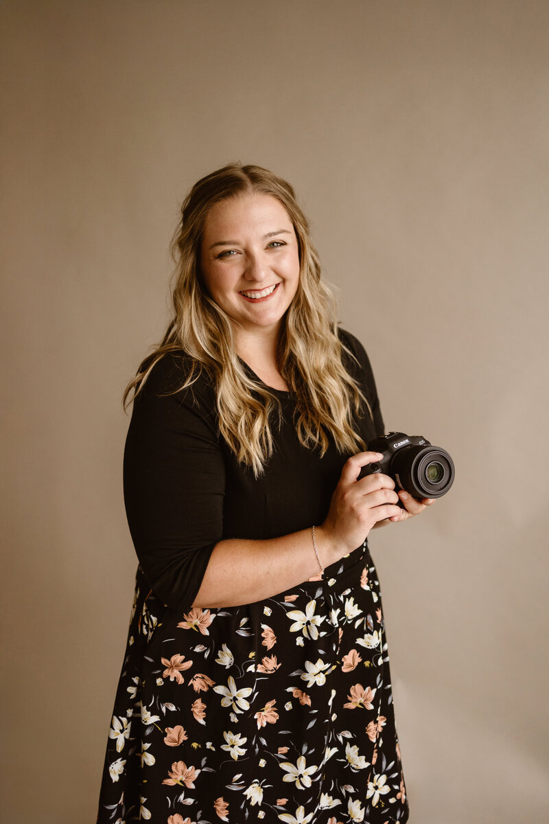 Cassandra, a smiling Denver-based photographer, holds her Canon camera while wearing a black floral dress in front of a neutral backdrop.