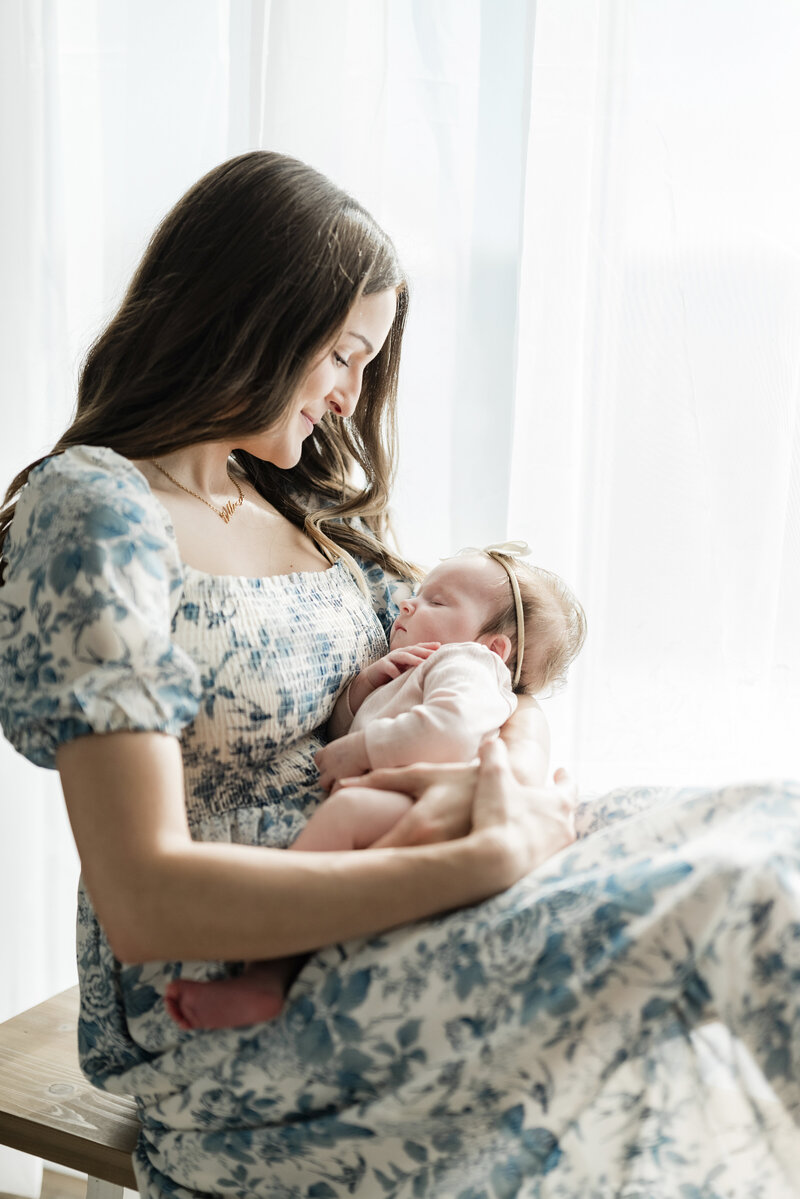 Mother in a floral dress holding her newborn baby near a softly lit window during a Kansas City newborn photography session.