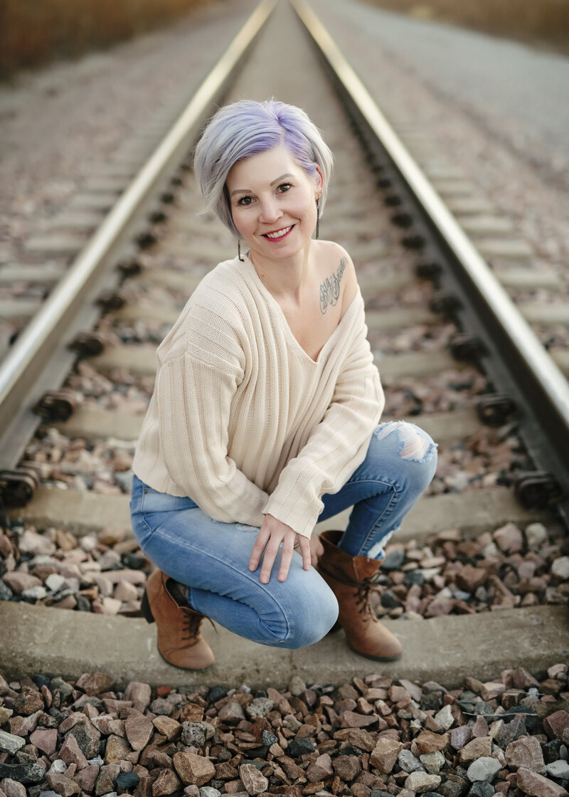 Brieanna Lebo kneeling on train tracks in a cream sweater and jeans, smiling in rustic outdoor photo.