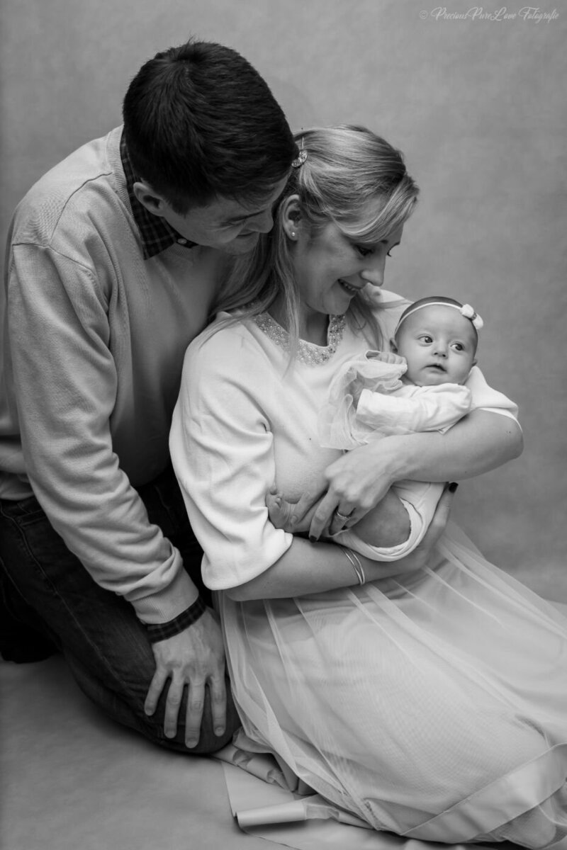 A black and white, posed family portrait of a mother, father, and their newborn baby. The mother is seated, holding the baby, while the father kneels next to them, both looking at the baby with love. The baby, wearing a white bow. A beautiful family photo of the mom holding their precious baby girl while the dad is admiringly looking to her.