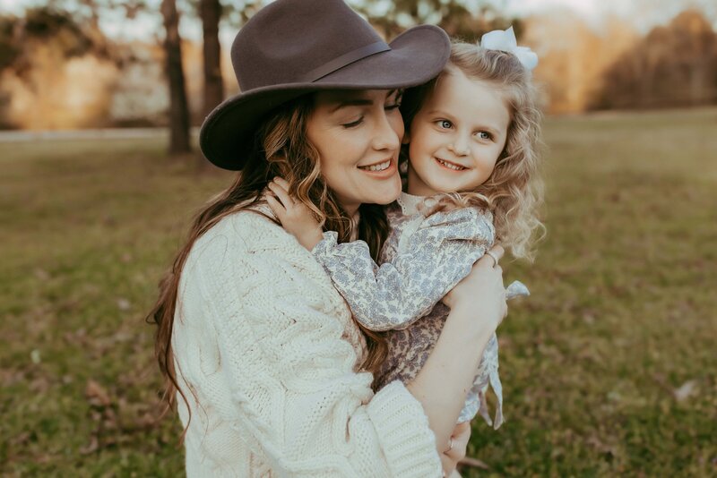In a grassy park, a woman in a hat and sweater warmly embraces her smiling, curly-haired child—a heartwarming moment beautifully captured by a Macon family photographer.