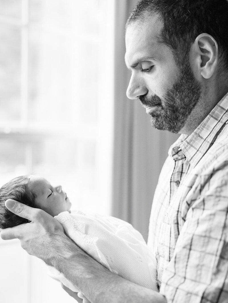 A father holding and looking down at his newborn while standing by a window by Katie Stansfield Photography, a Richmond newborn photographer.