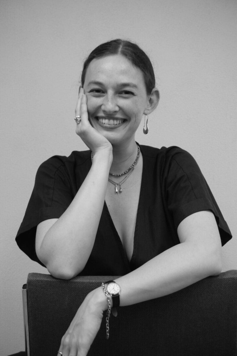 Cora Demary smiling  in a black shirt resting her chin on her hand.