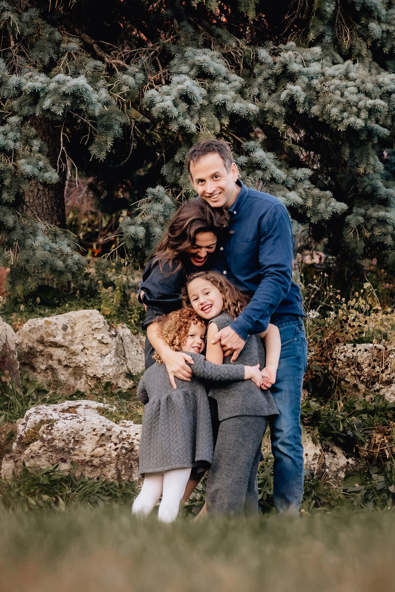 A family of four huddled together in front of a tree, smiling and embracing warmly, with the parents leaning down to hug their two young daughters who are grinning joyfully.