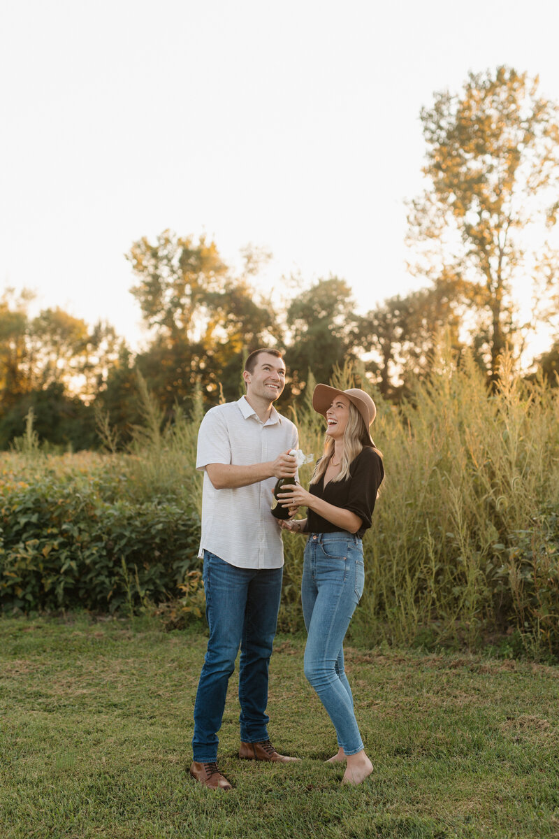 Man and woman in grassy field popping a bottle of champagne during their engagement photos