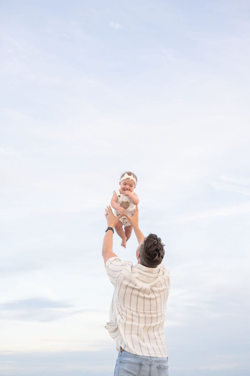 father gently tosses smiling baby girl into the sky as he reaches up to catch her