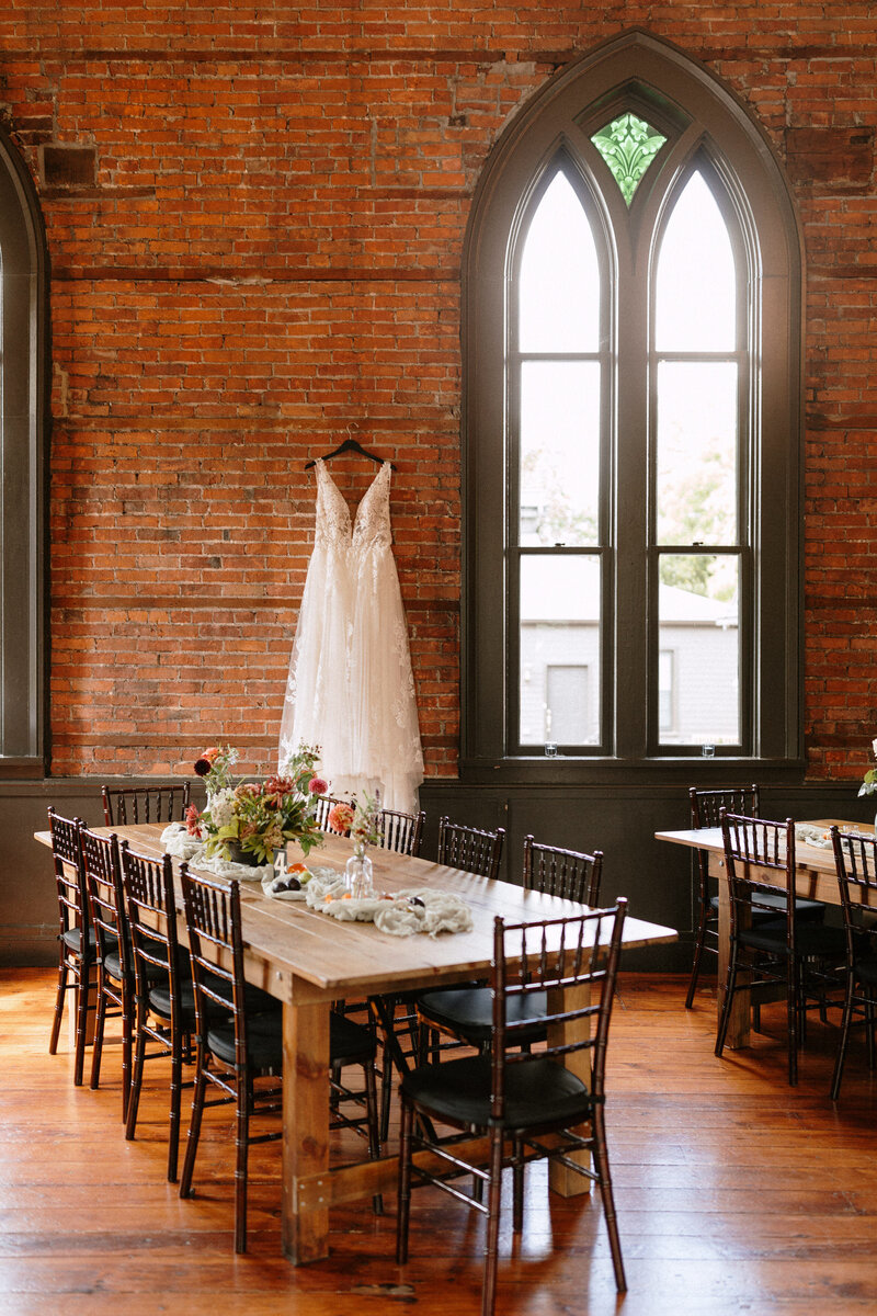 A bridal gown hangs against a rustic brick wall, with elegantly set tables adorned with floral centerpieces in a charming venue.