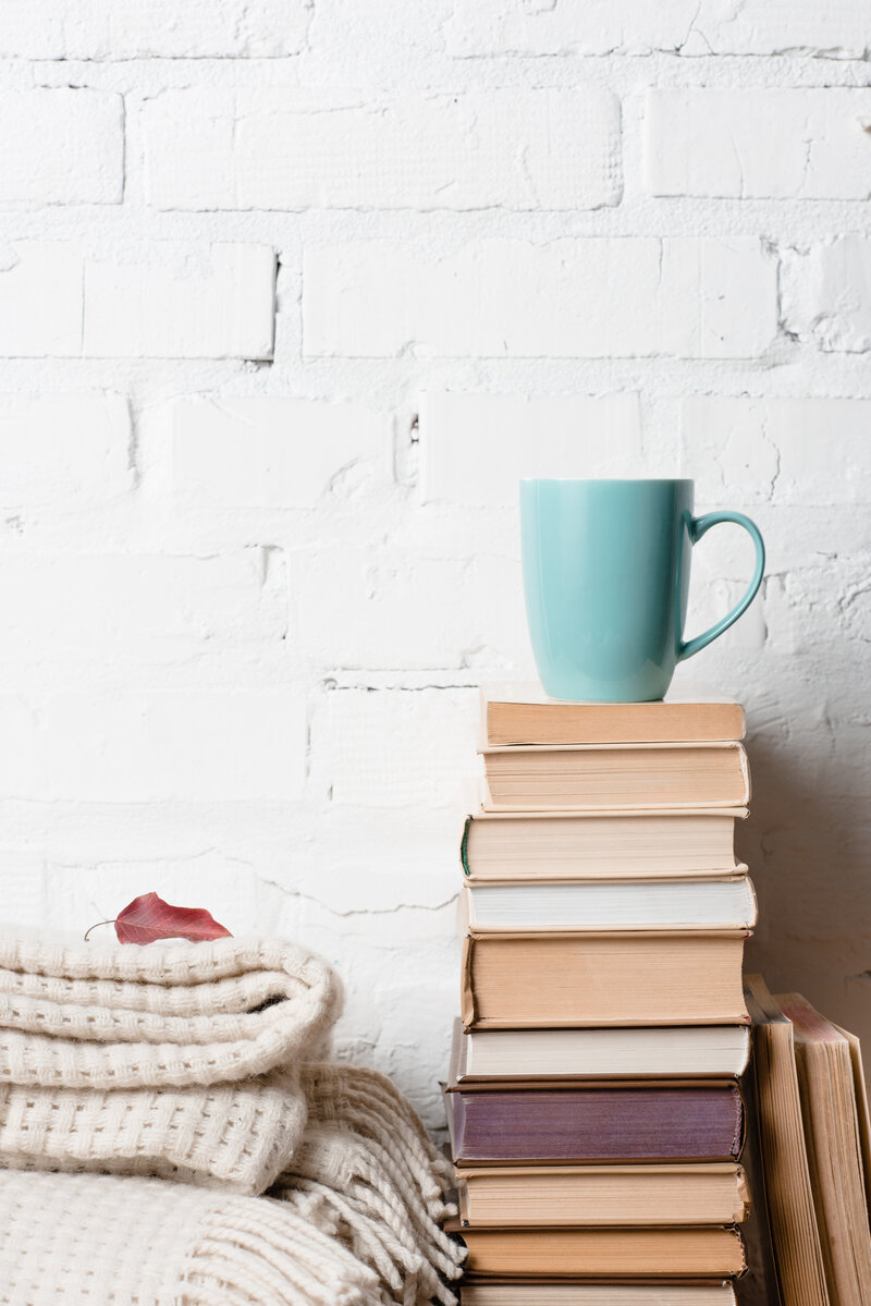 Stack of books with a teal mug on top, next to folded cozy blankets, against a white brick wall background—warm and inviting reading or blogging setup.