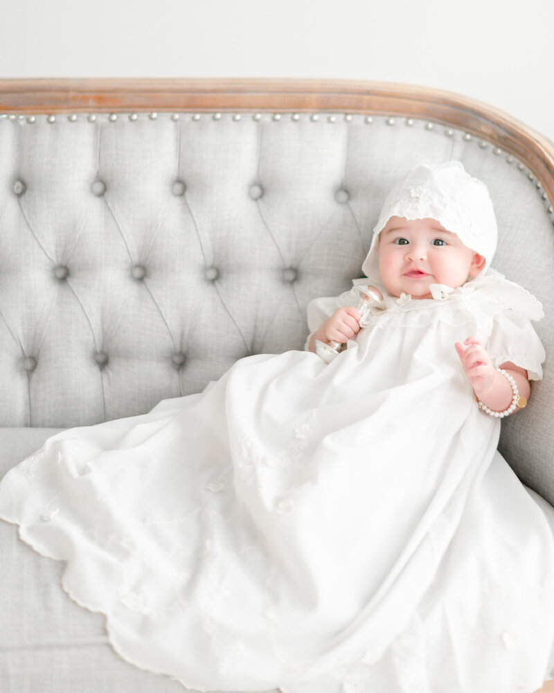 3 month old baby in baptism gown holds a silver rattle and wears a bonnet