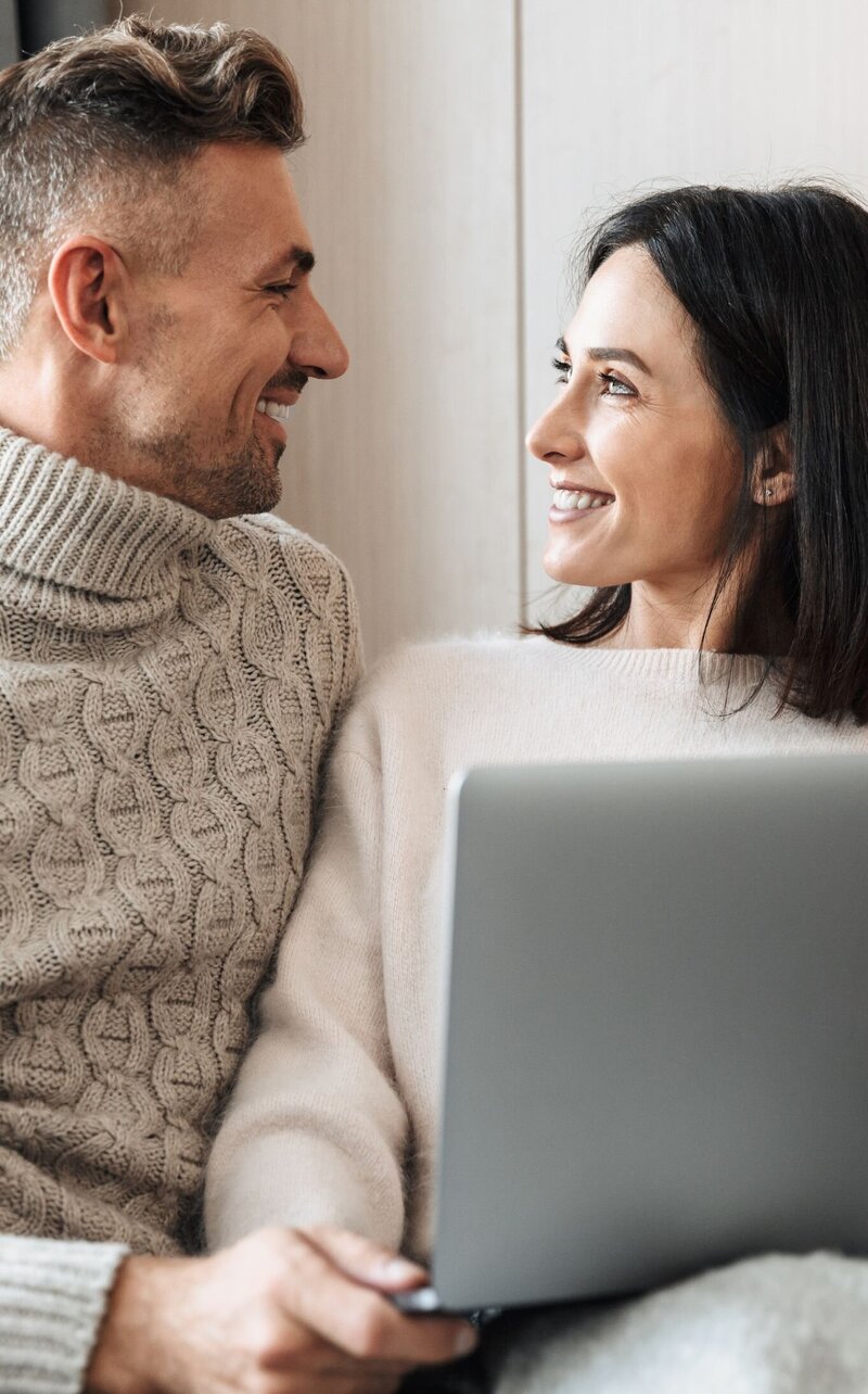 A couple looking and smiling at each other while holding a laptop. This can represent a couple in the aftermath of infidelity feeinlg accomplished for transforming pain into strength with our expert-led program.