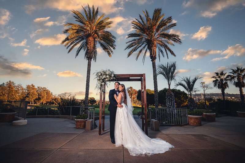 Newlywed bride and groom posing at their wedding arch, staged before the sprawling golf course and towering palm trees, after their wedding ceremony at Talega Golf Club.