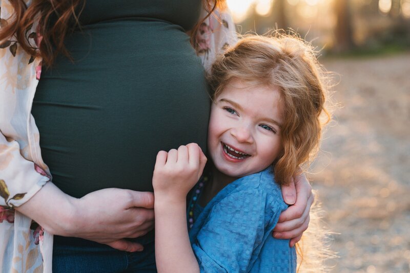 Preschool aged girl smiling and hugging mom's round, pregnant blly