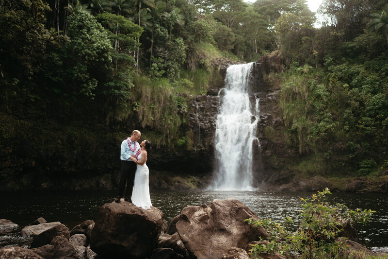 Eloping ideas | Eloping couple stands on top of a huge rock with a big waterfall behind them to the right