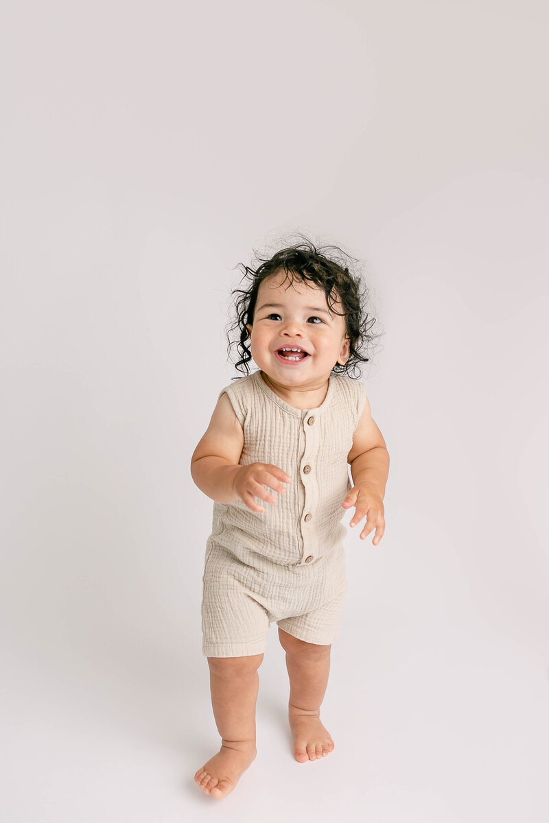 One Year old baby standing and looking a bit off camera. Wearing a beige neutral romper at portland portrait photography studio session.