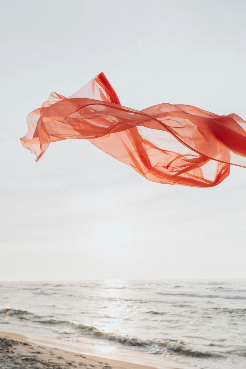 Vibrant orange fabric billowing in the wind near a tranquil body of water, creating a sense of movement and serenity against the natural ocean backdrop.