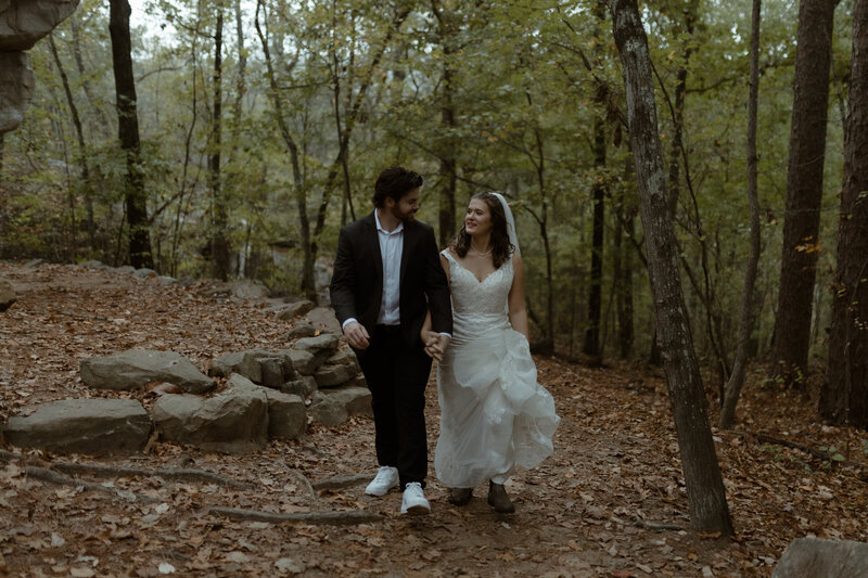 bride in lace wedding dress and groom in suit walking hand-in-hand through a forest trail during a fall adventure elopement