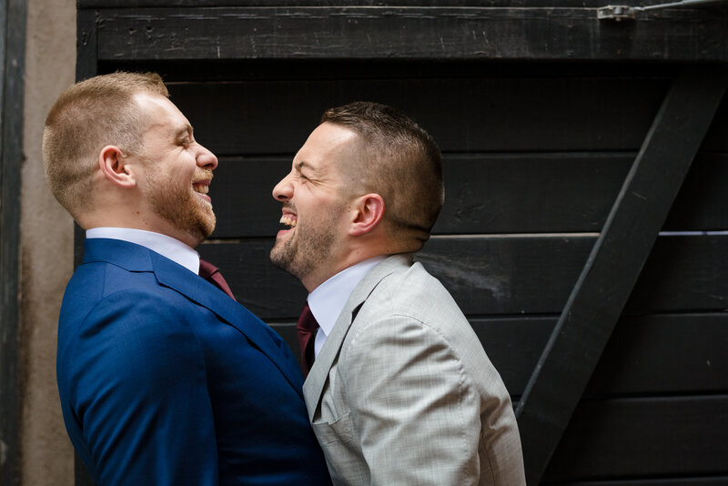 Two grooms wearing suits and laughing in front of a black door on their wedding day at dye house providence ri