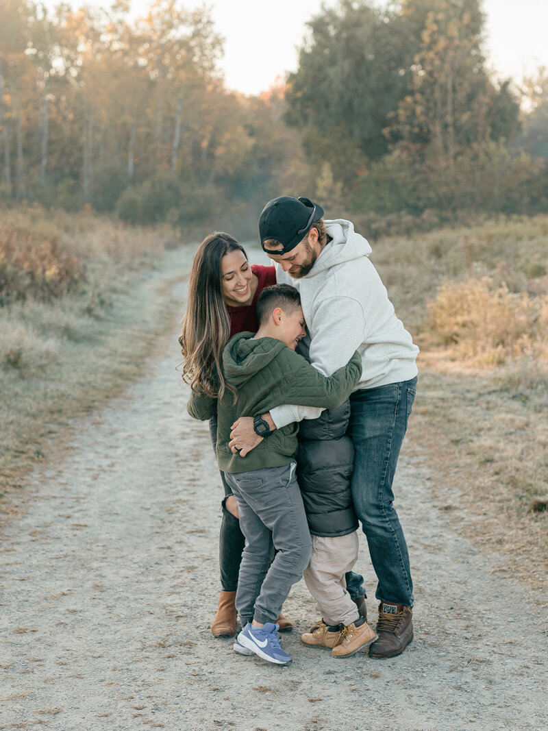 A couple shares a warm embrace on a scenic outdoor path, surrounded by soft golden light. Their connection is evident in their joyful expressions, creating a timeless and heartfelt moment captured in nature. Photographed by KCP Families.