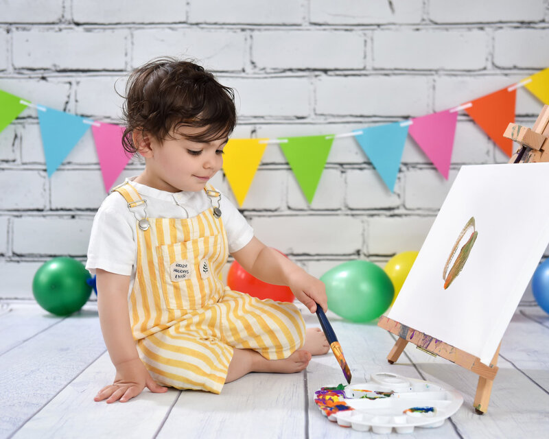 A joyful toddler in yellow striped overalls holding large "TWO" letters during a vibrant two-year-old milestone photo shoot on a white wooden floor.