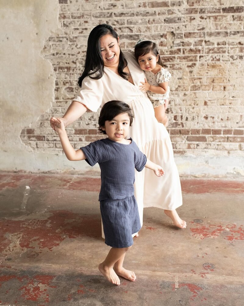 Mother holding a toddler and dancing with her barefoot son in front of a rustic brick wall.