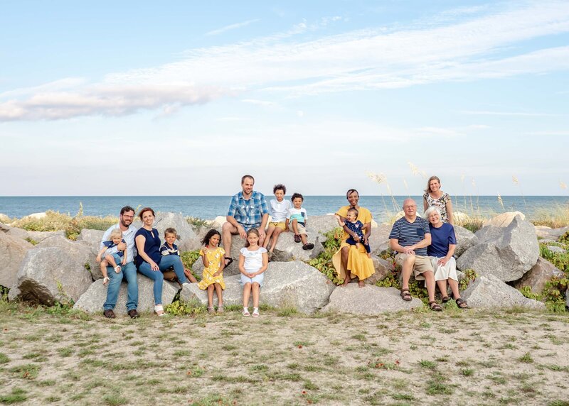 A large family sits on rocks by the ocean at Fort Fisher, enjoying a sunny day. The group includes multiple generations, dressed casually, with children playing and adults smiling. This image is perfect for destination family photographer NC, capturing the joy of family reunions.