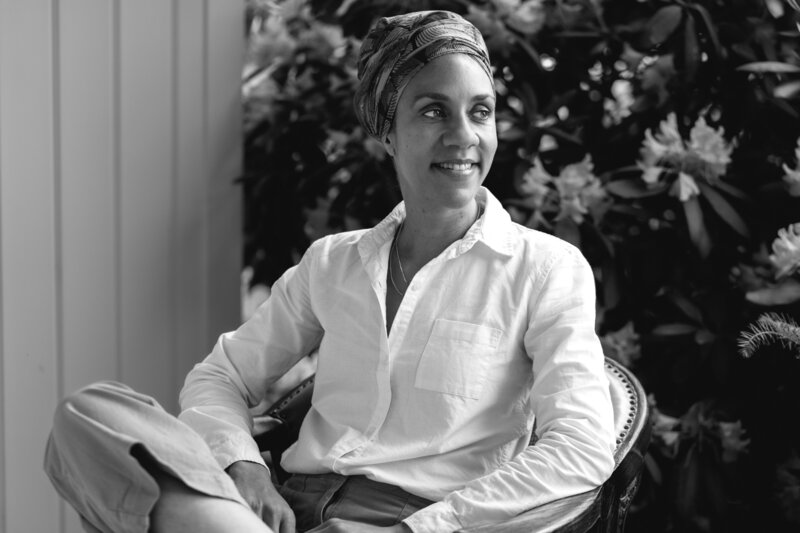 A Black comics artist and author sits in a chair in front of large flowers. She's wearing a headwrap and looking to the side, smiling.