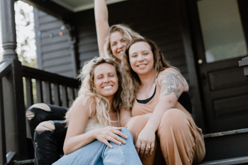 Three women sit closely together on outdoor steps, smiling and looking relaxed. One raises her arm in the background. The casual, friendly setting with a dark wooden house behind them hints at a break during a white label design course.