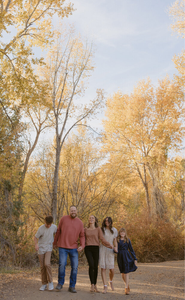 A husband and wife holding hands with their 2 daughters and son, smiling and walking towards the camera. They are outside with trees around them during Fall.