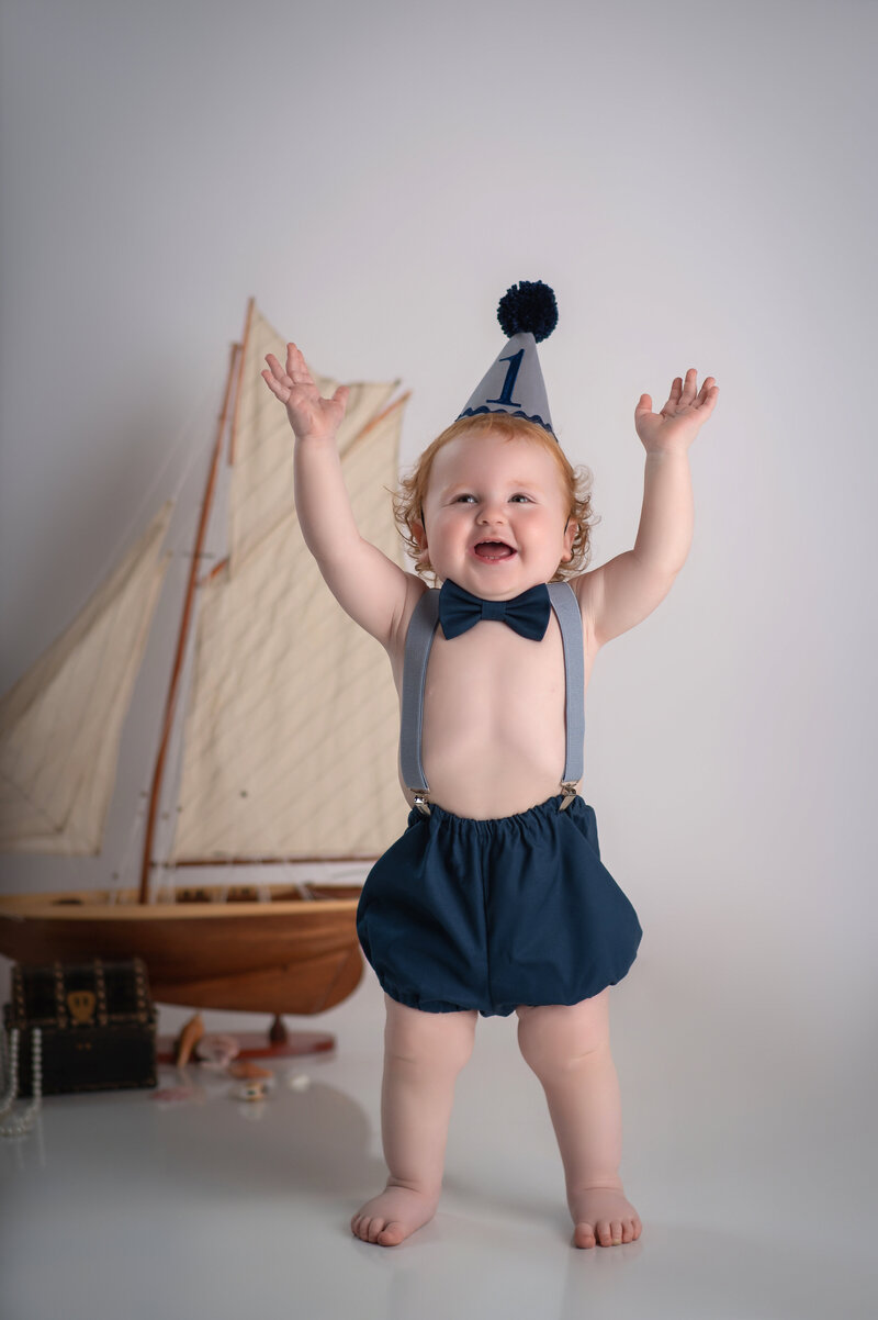 milestone portrait for one year old boy with red curly hear wearing suspenders and navy blue bloomers and a pointy number one birthday hat with his arms up in the air and smiling in front of a sailboat prop and white backdrop