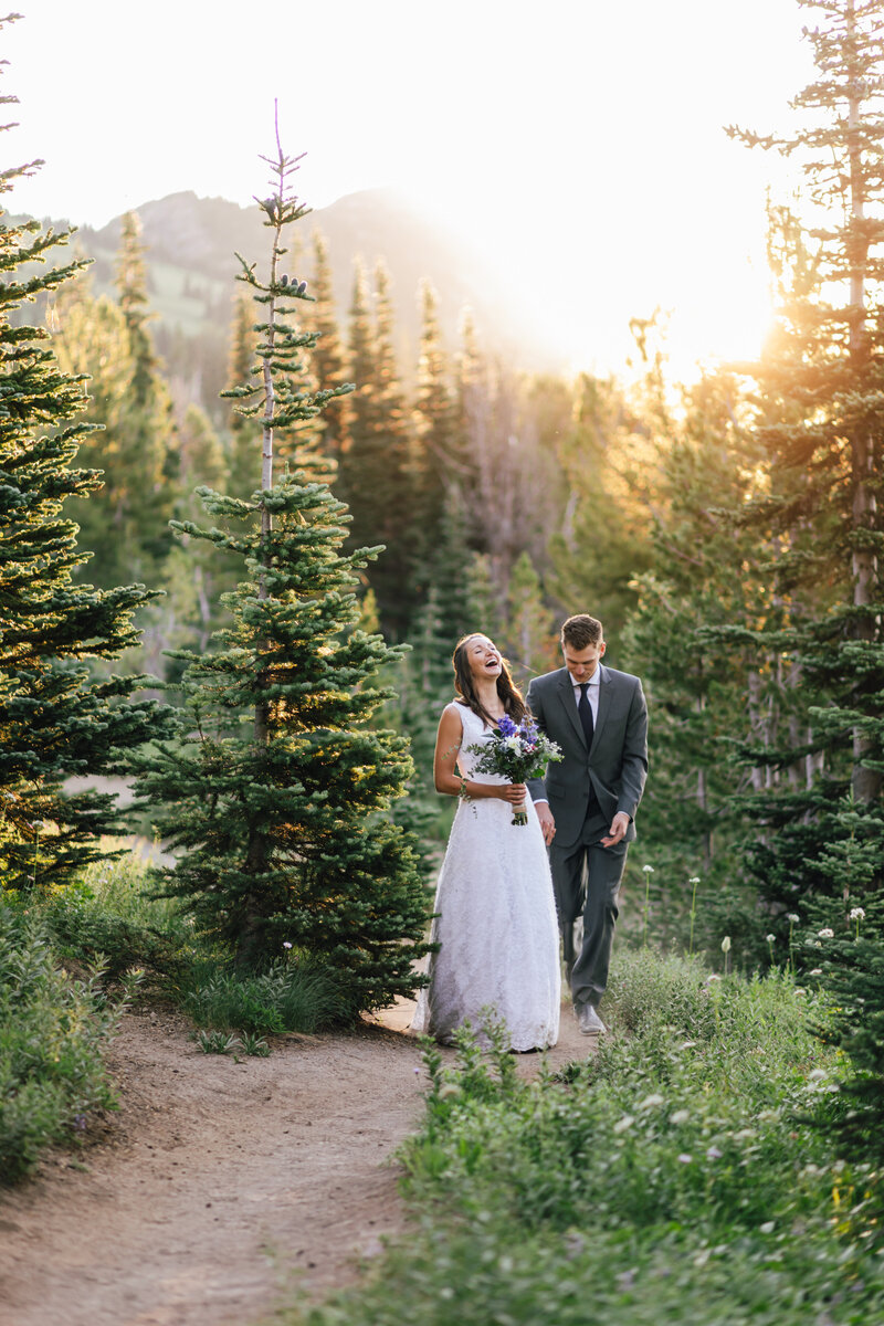 A bride, with her hair down wearing a white lace gown throws her head back in laughter holding a bouquet of wildlfowers in one hand and her groom's hand in another as they hike a trail at sunrise on their elopement day at Mt Rainier National Park in Washington. | Erica Swantek Photography