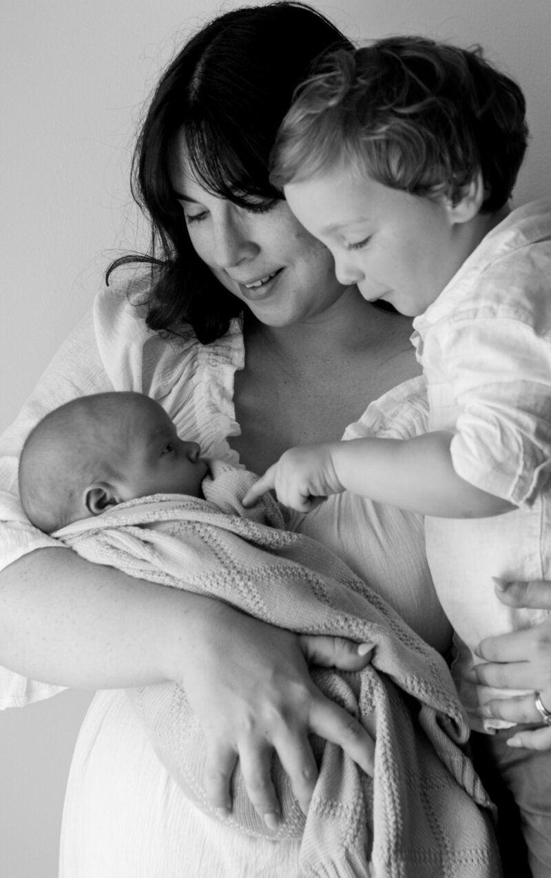 Black and white photo of a mother cradling her newborn while an older sibling gently reaches out to touch the baby, capturing a quiet and emotional family moment.