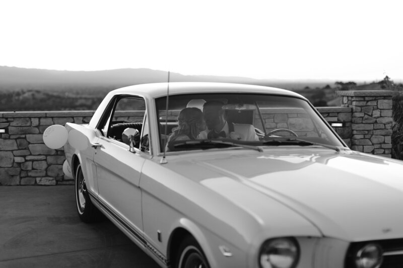 Bride and groom kissing in front seat of white vintage getaway car