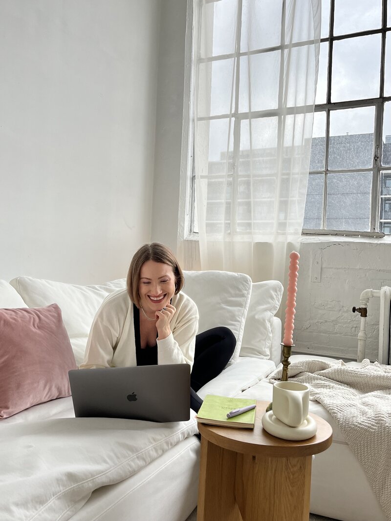 Margarita, a copywriter for websites, sitting on a white couch in a loft with big windows behind her, smiling at her laptop in front of her; a notebook, white coffee mug, and pink squiggly candle is on the small coffee table in front of her.