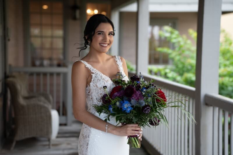 Smiling bride in white lace wedding dress holding a burgundy, purple and blue floral bouquet, standing on a covered porch with white railings at the White Room venue in historic downtown St. Augustine