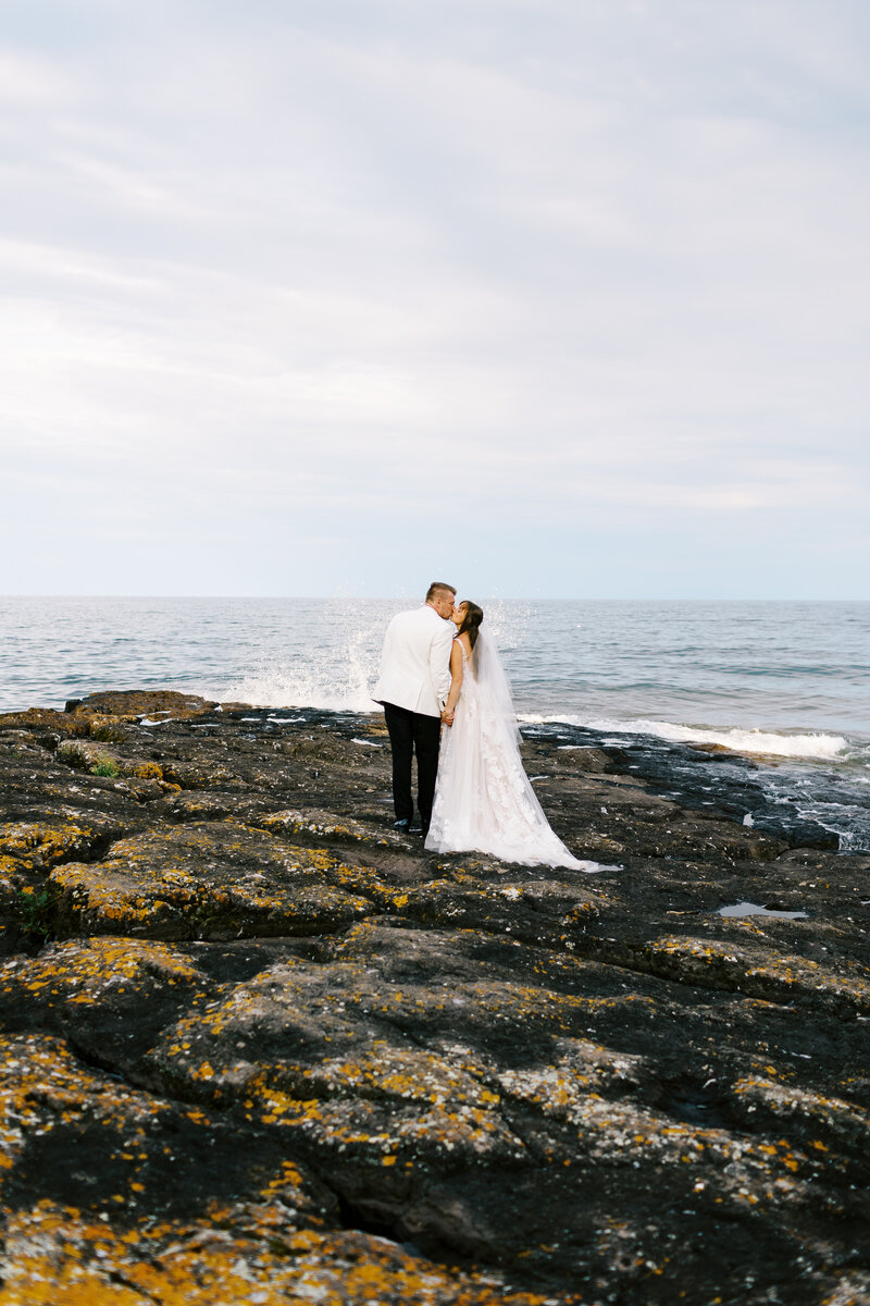 Bride and groom share a romantic kiss on the rocky shoreline of North Shore, with the vast expanse of Lake Superior in the background. Captured by Toly Dzyuba Photography, Minneapolis wedding photographer.