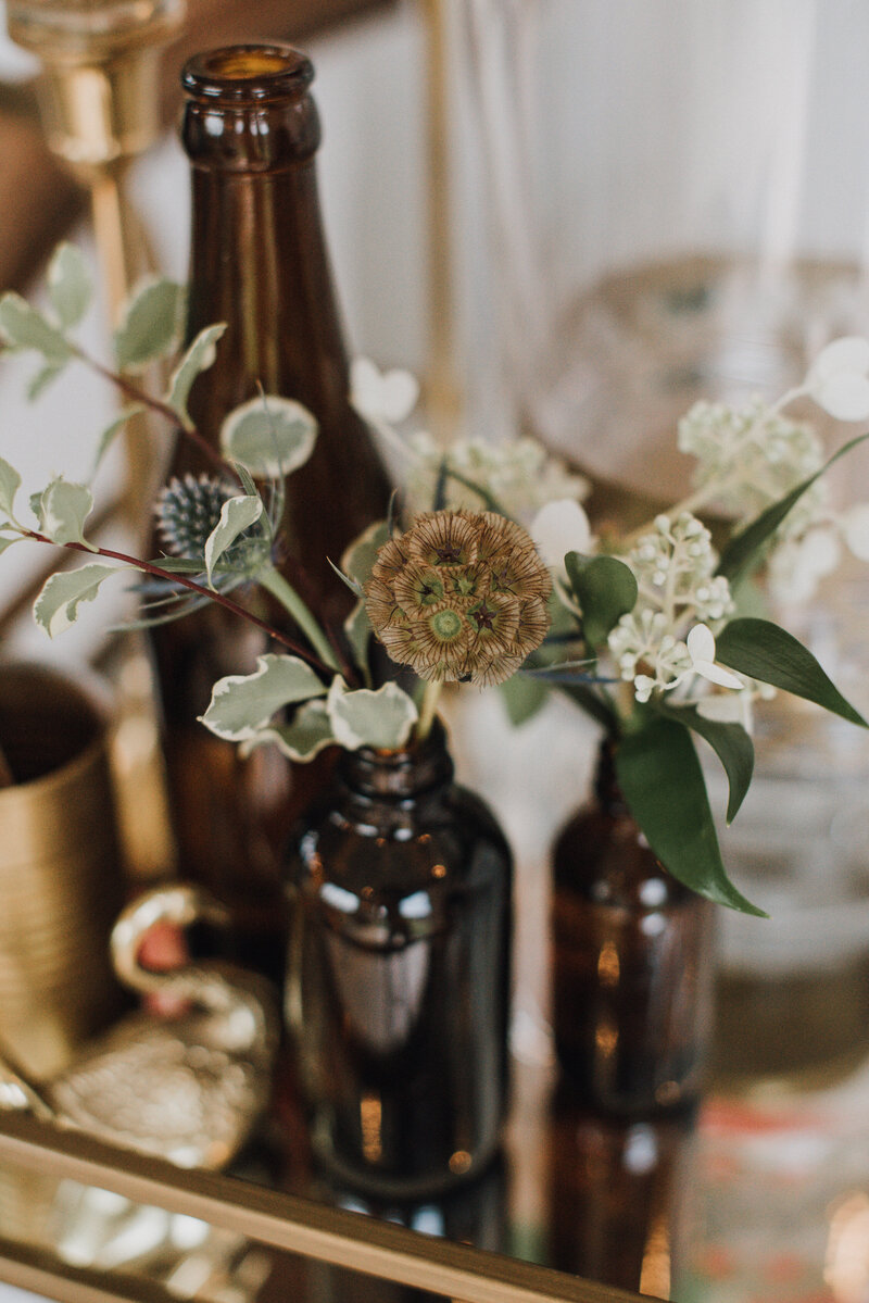 Amber glass bud vases on a brass midcentury bar cart hold sprigs of scabiosa, thistle, and hydrangea