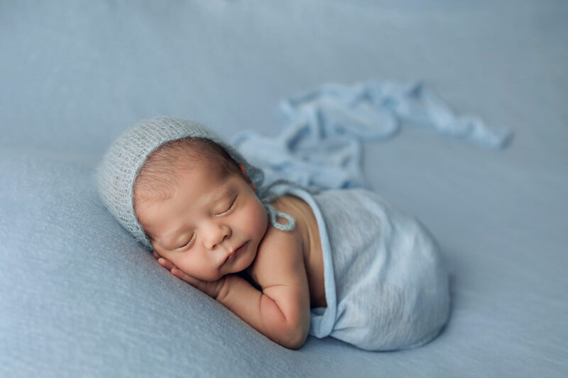 A newborn baby is peacefully sleeping, curled up on a soft, dusty rose-colored background. The baby is naked, showcasing their delicate, natural form with adorable baby rolls. A floral headband with pink and green flowers adorns the baby's head, adding a touch of sweetness to the serene scene. The baby's tiny hands are gently placed near their face, and their expression is calm and content, creating a heartwarming and tender portrait.