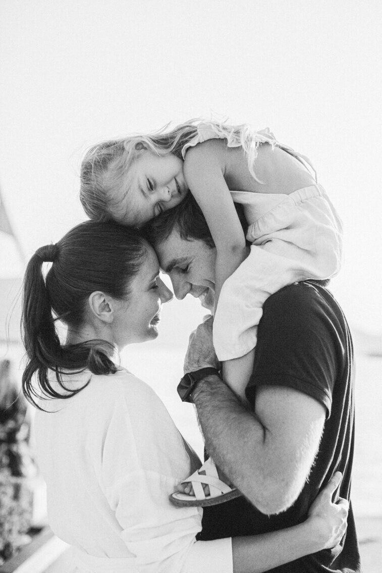 Black and white photo of parents hugging with their smiling daughter on dad’s shoulders, captured during a candid family photoshoot in Toronto