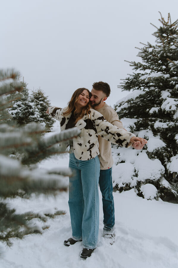Standing behind his girlfriend, a man holds her hands. They're stand in the middle of a snowy forest for a fun couples adventure session in Idaho Falls, ID.