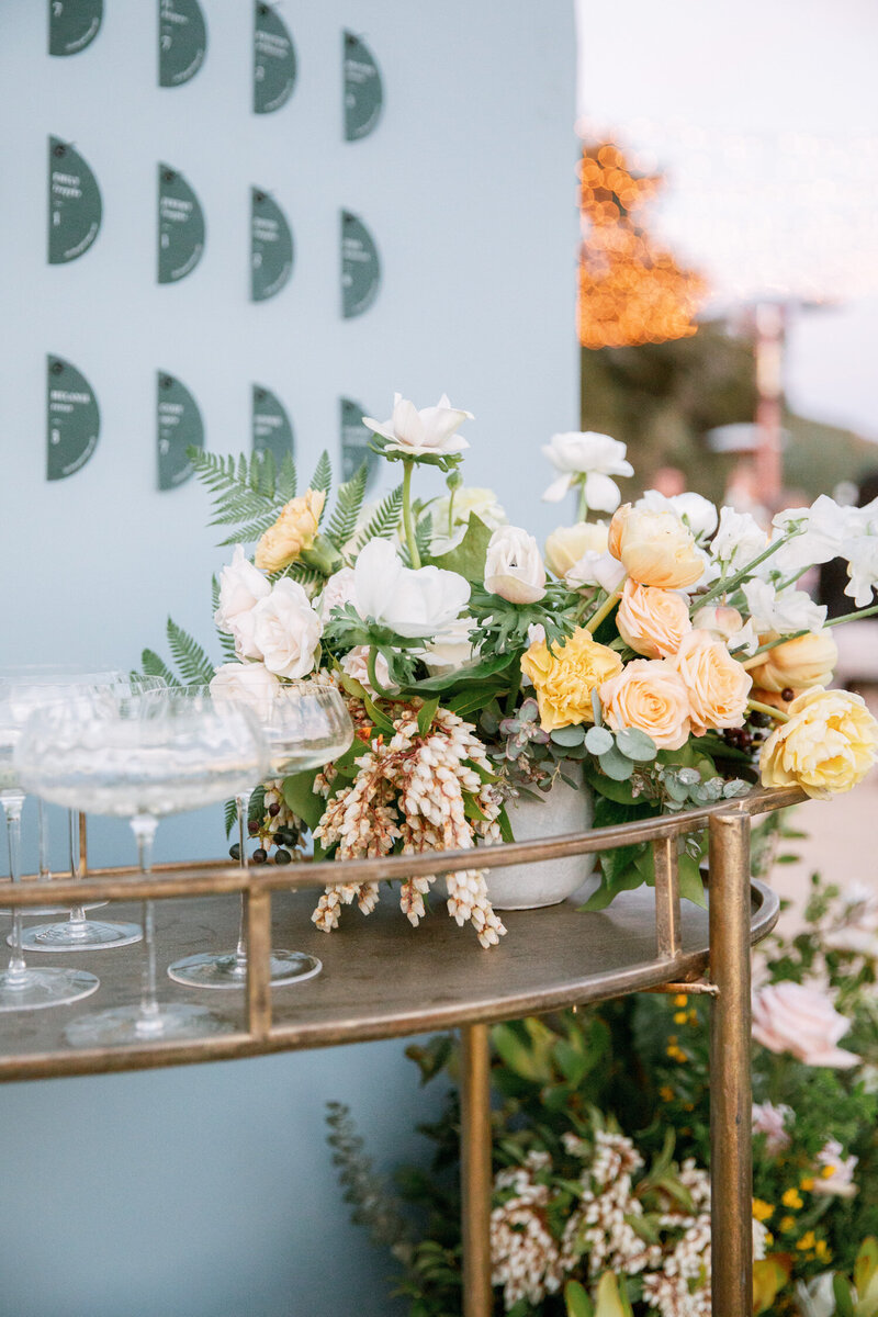 Gold cocktail cart with champagne coupes on it and a floral centerpiece