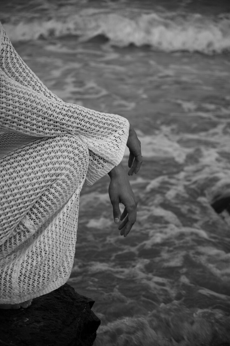 Black and white photo of a woman on a cliff. Up close perspective of only her hands and the side of her legs. The sea in the background