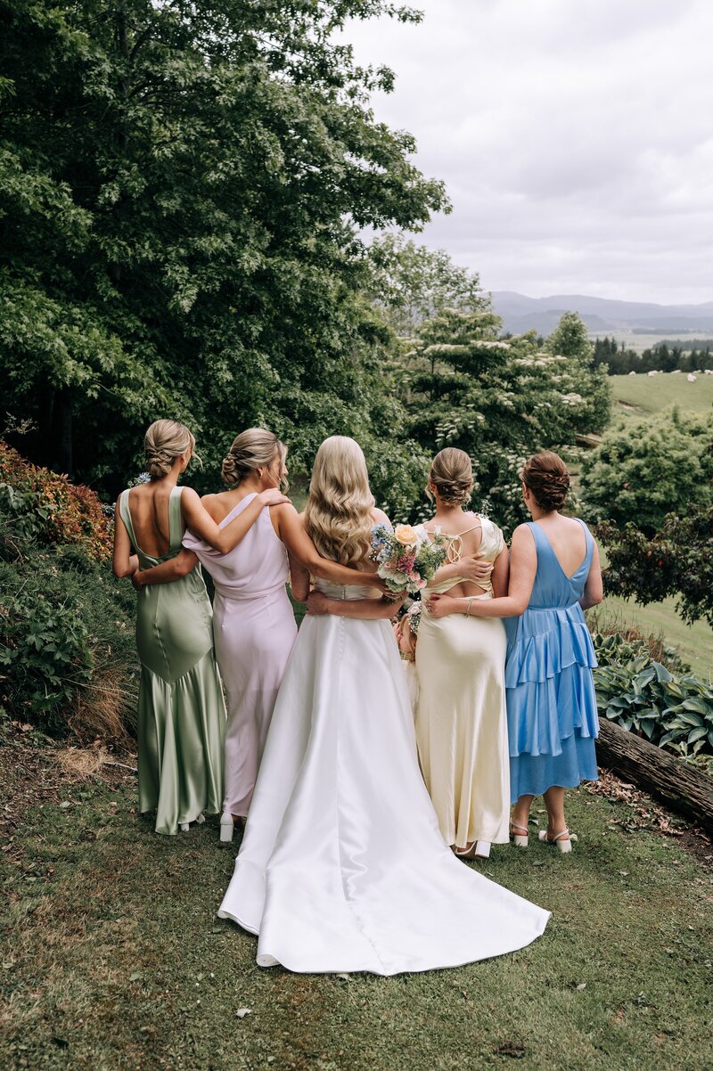 the backs of bridesmaids in mismatched pastel dresses overlooking green waimumu fields at wedding