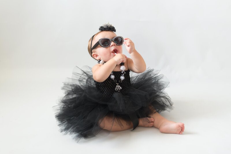 little girl as audrey hepburn in black tutu, necklace, sunglasses and eating a lollipop
