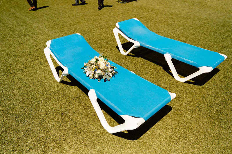 Two empty blue lounge chairs sit on green grass, a white and green floral bouquet atop one chair. Shadows and guests’ legs in the background hint at a celebration—perfectly captured by a film photographer nj for timeless wedding memories.
