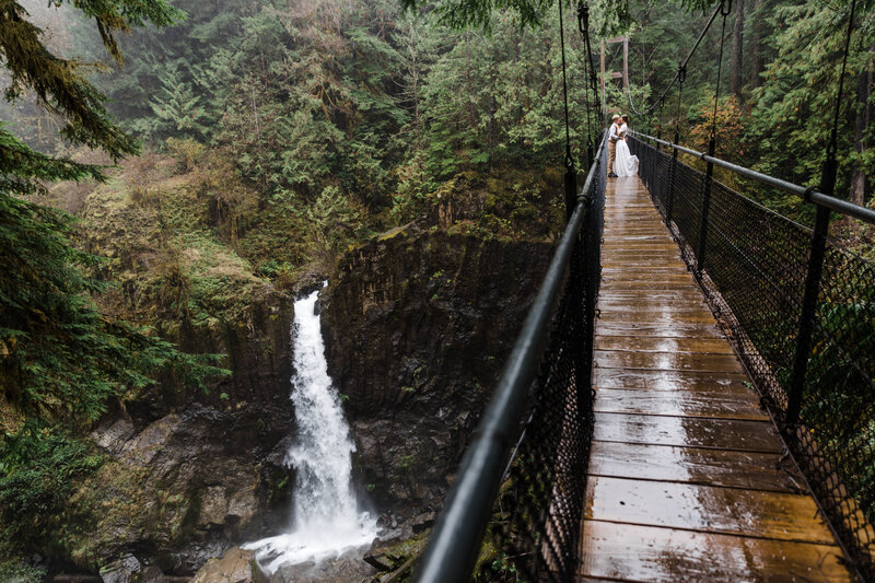 A LGBTQ+ couple embraces on a wet suspension bridge over Drift Creek Falls at their rainy Oregon elopement. 