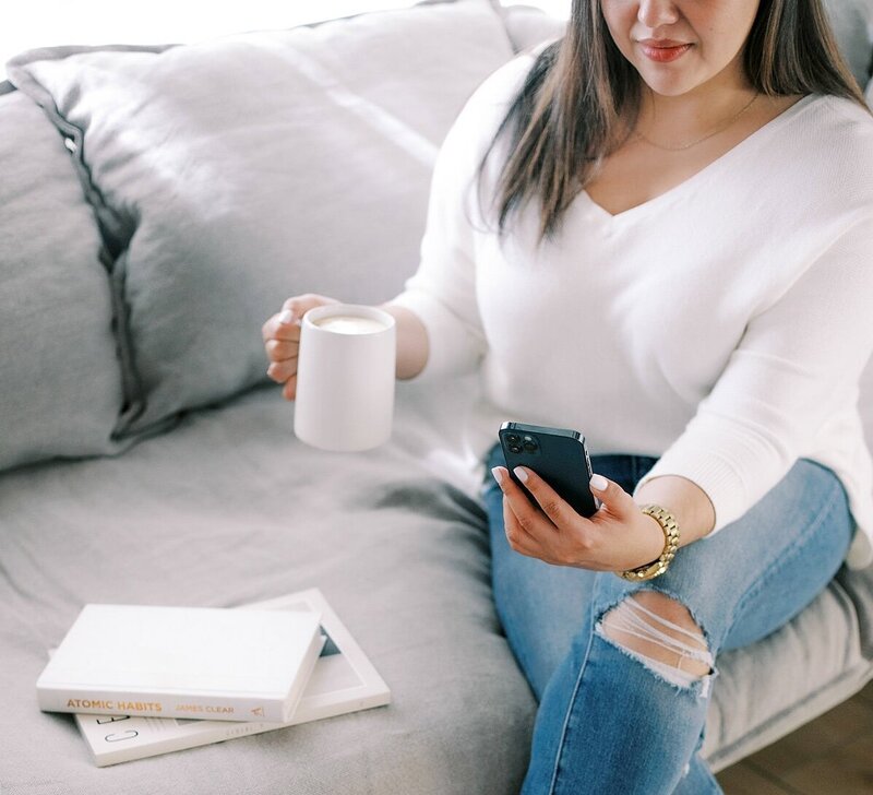 Woman in a white sweater and ripped jeans sitting on a gray sofa, holding a coffee mug and smartphone with books beside her.